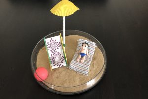 there is sand in a glass bowl and in it a small doll's deckchair and a small figure made of modelling clay lying on a tiny towel, next to it a small ball and a small parasol