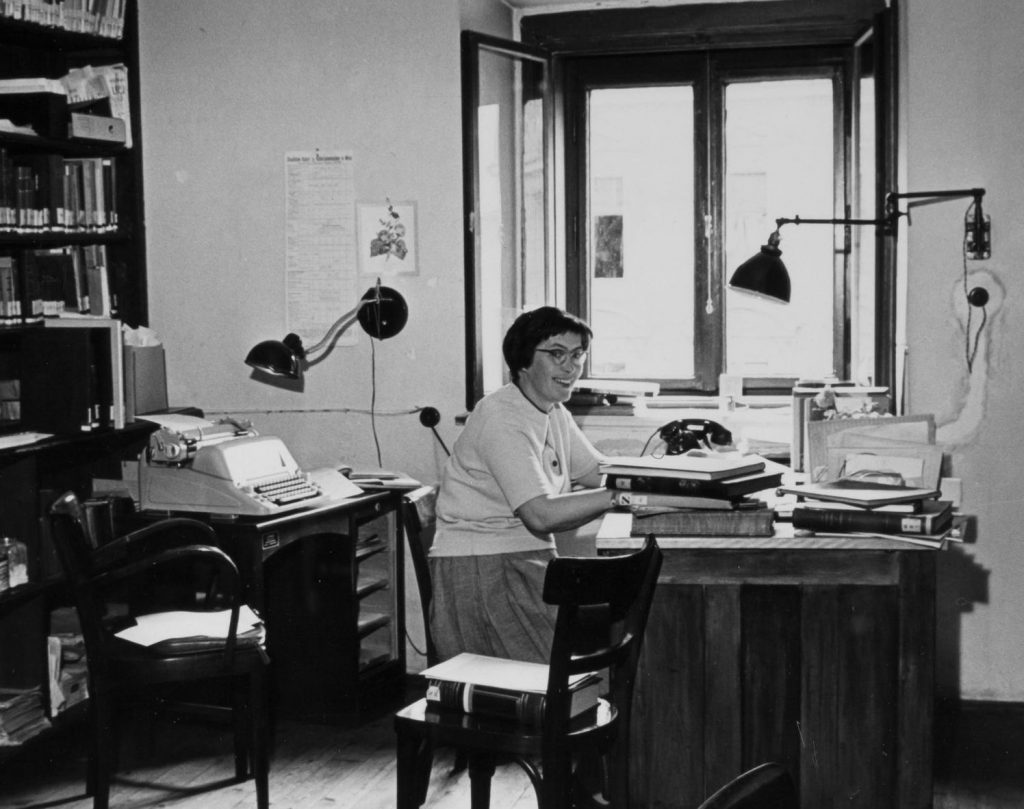 schwarz-weiß Foto mit Frau an einem Schreibtisch, sie lächelt und trägt Brillen