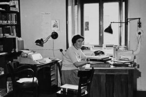 black and white photo with woman at a desk, she smiles and wears glasses