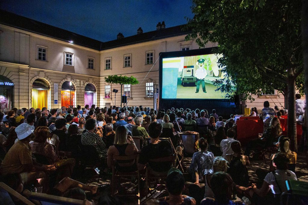 a lot of people in a courtyard sitting in front of a screen at dusk