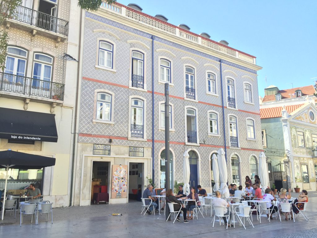 multi-storey house with patterned tiles, white chairs with sunshades stand in front of it on a square