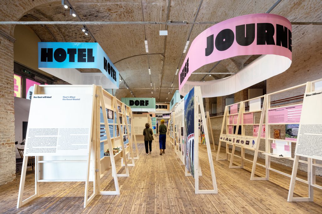 Ausstellungsraum mit 2 Personen, darüber die Schilder mit Schrift 