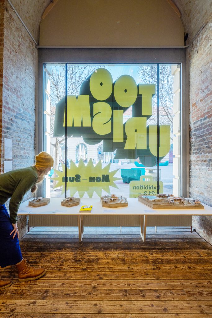 Person with yellow bonnet looking at exhibition objects, behind them large upside-down lettering