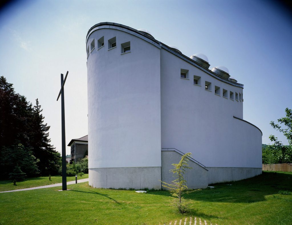 rund gebautes Gebäude in einer Wiese stehend, davor steht ein großes Kreuz