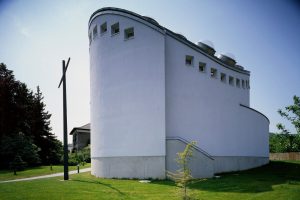 rund gebautes Gebäude in einer Wiese stehend, davor steht ein großes Kreuz