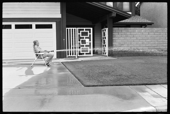 b/w photo of a man in an armchair spraying a meadow with a garden hose