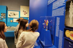 Four girls stand in front of a blue wall with texts and two Barbie dolls on display