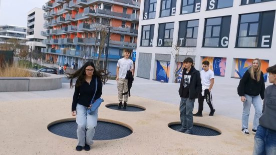 Sechs Jugendliche vor Wohngebäuden, ein paar davon hüpfen auf Trampolinen