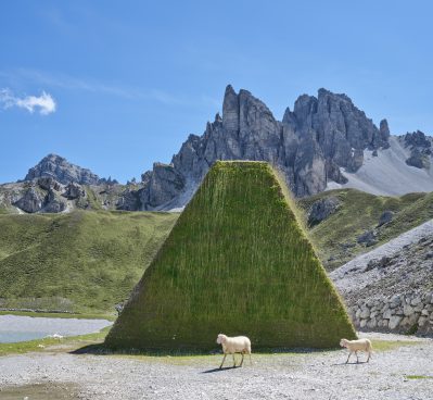 Hochgebirge im Hintergrund, davor Schafe vor einem pyramidenartigen Turm aus Gras