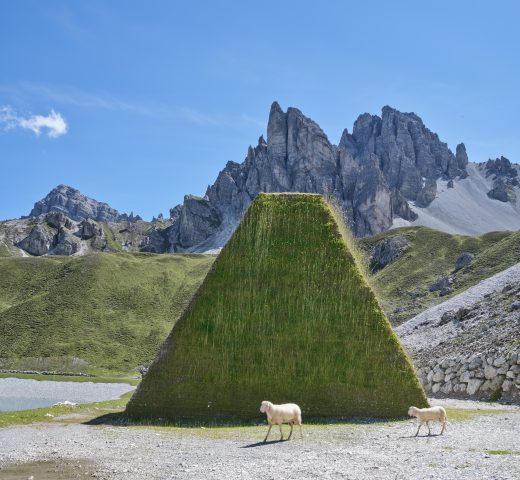High mountains in the background, sheep in front of a pyramid-like tower of grass