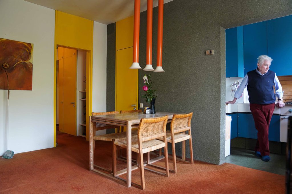 Mann mit weißen Haaren steht in einer blauen Küche, im Vordergrund das Esszimmer mit Holzsesseln und roten Röhrenlampen, die von der Decke hängen