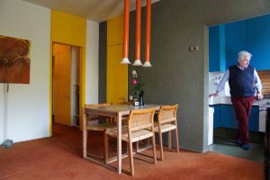Mann mit weißen Haaren steht in einer blauen Küche, im Vordergrund das Esszimmer mit Holzsesseln und roten Röhrenlampen, die von der Decke hängen