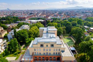 Ansicht eines großes Gebäudes von oben, Farbe ist beige und im Hintergrund sieht man viele Häuser und Bäume
