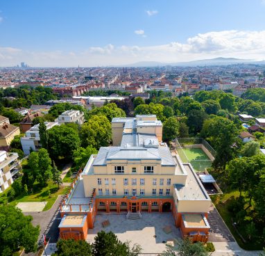Ansicht eines großes Gebäudes von oben, Farbe ist beige und im Hintergrund sieht man viele Häuser und Bäume