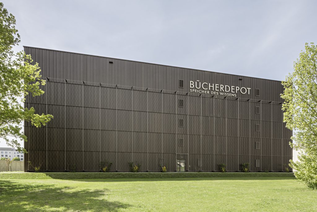 graues Gebäude, das auf einer Wiese steht. Auf dem Gebäude steht Bücherdepot Speicher des Wissens