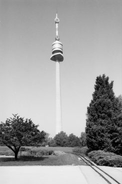 Black-and-white photo showing a tall tower with a road and railroad tracks in front of it