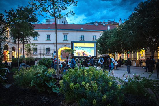Courtyard with plants in the foreground, large screen in the background with many people sitting in front of it