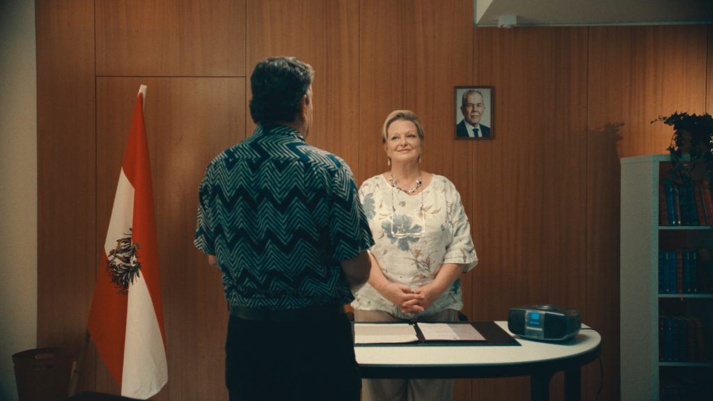 Frau steht vor Holzwand, neben ihr eine Fahne mit einem Wappen und die Farben rot-weiß-rot. Mit dem Rücken zur Kamera steht der Frau zugewandt ein Mann.