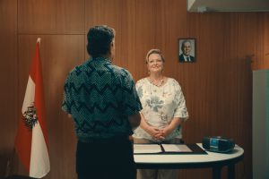 Frau steht vor Holzwand, neben ihr eine Fahne mit einem Wappen und die Farben rot-weiß-rot. Mit dem Rücken zur Kamera steht der Frau zugewandt ein Mann.