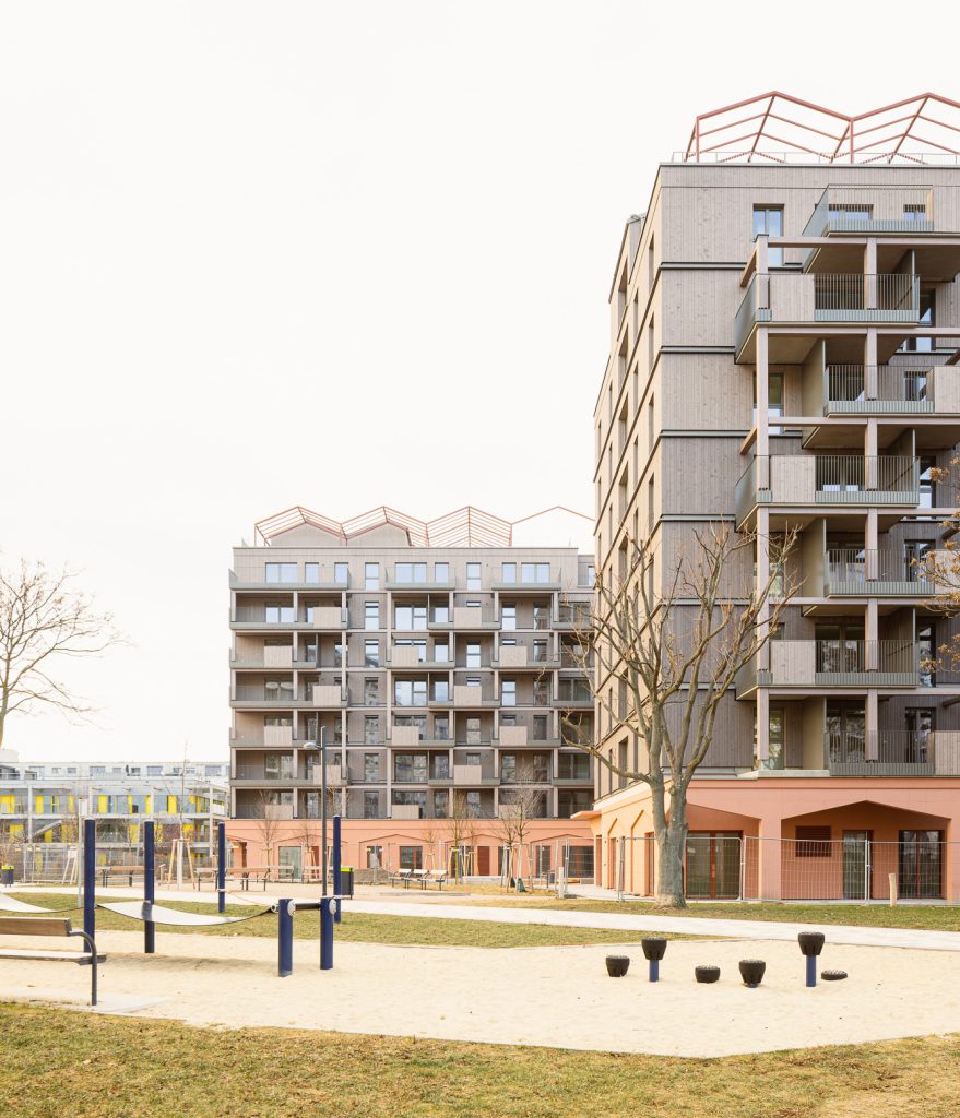zwei grau-rosa mehrgeschossige Gebäude, davor ist ein Spielplatz zu sehen