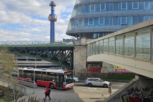 Straße mit oberhalb zwei Brücken, eine davon verglast. Dahinter ein hoher Trum mit einem runden Element im oberen Drittel