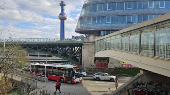 A street with two bridges above it, one of which is glass-covered. Behind it, a tall structure with a circular element in its upper third