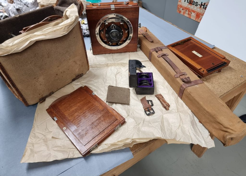 auf einem Tischen liegen mehrere Gegenstände, darunter eine sehr alt aussehende Kamera, eine abgerissene Gürtelschnalle etc.