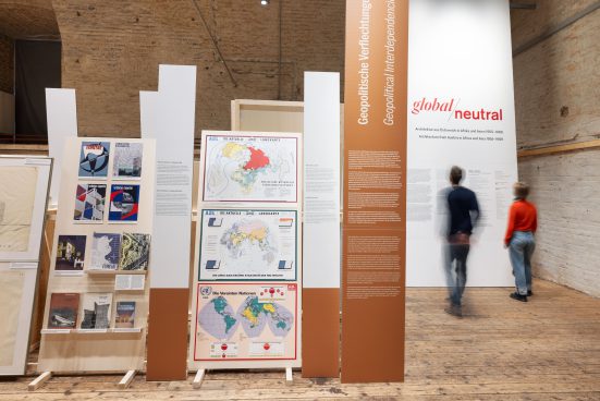 Two people are standing in front of a very high wall bearing the words “Global / Neutral”. In the foreground, there are pictures and information panels.