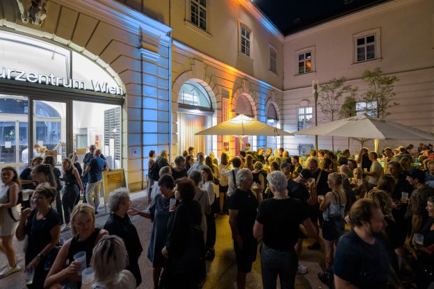 Many people are standing in the dark in a lit courtyard. In the background are two large umbrellas.