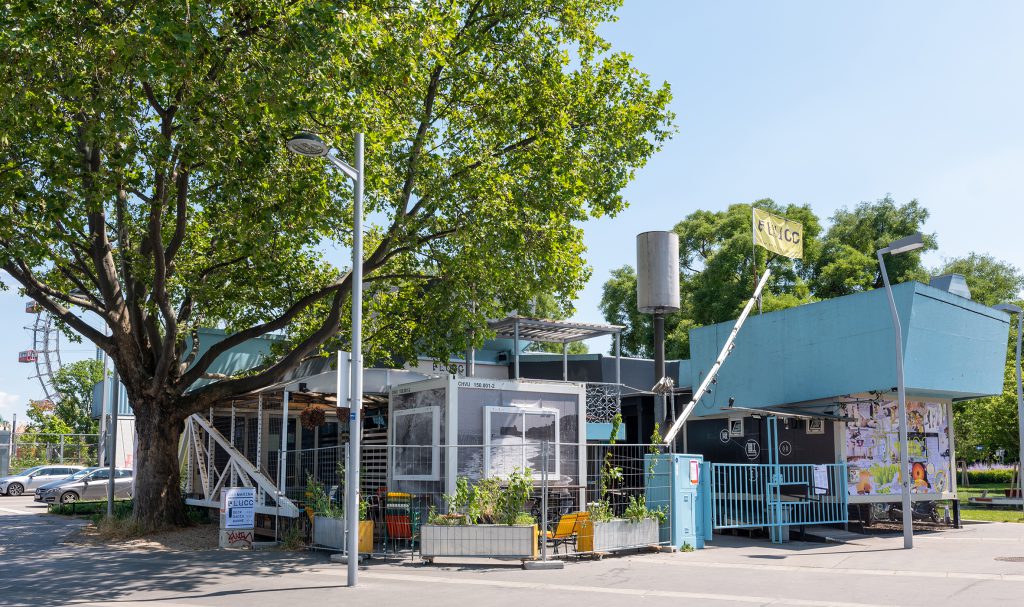 ein Gebäude mit hellblauem Dach und Gitter. Es ist mit einem Bauzaun eingezäunt. Im Hintergrund sieht man das Riesenrad.