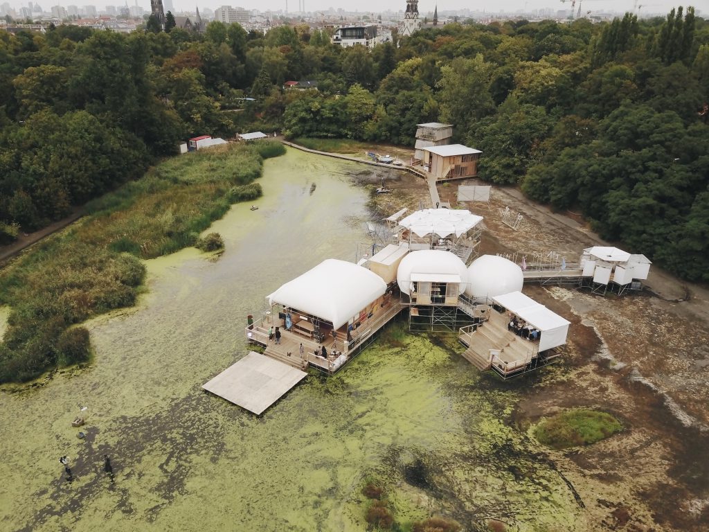 Blick von oben auf einen Teich im Wald, worin sich eine Art Haus bzw mehrere Häuser befinden
