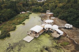 Blick von oben auf einen Teich im Wald, worin sich eine Art Haus bzw mehrere Häuser befinden