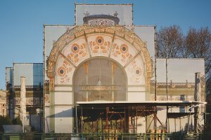 Ein Gebäude mit einem Rundbogen mit sehr viel Verzierungen in Form von Ornamenten wie Blumen, Blättern... meistens in Gold, aber auch die Farbe Grün ist zu sehen. In der Mitte steht Karlsplatz. Davor ist ein Gerüst aufgebaut.