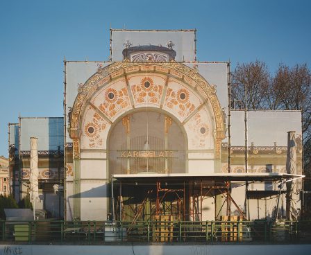 A building with a round arch featuring extensive decorations in the form of ornaments such as flowers and leaves... mostly in gold, but the color green is also visible. Karlsplatz is located in the center. Scaffolding has been set up in front of it.