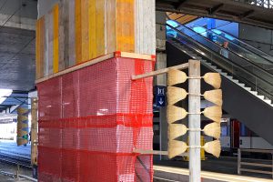 Man sieht Gleise, offenbar handelt es sich um einen Bahnhof. Zwischen den Gleisen ist ein Teil mit Holz und roten Lochplanen abgedeckt. Davor ist eine Konstruktion aus Holz, aus der seitlich kleine Besen rausragen.