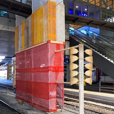 Man sieht Gleise, offenbar handelt es sich um einen Bahnhof. Zwischen den Gleisen ist ein Teil mit Holz und roten Lochplanen abgedeckt. Davor ist eine Konstruktion aus Holz, aus der seitlich kleine Besen rausragen.