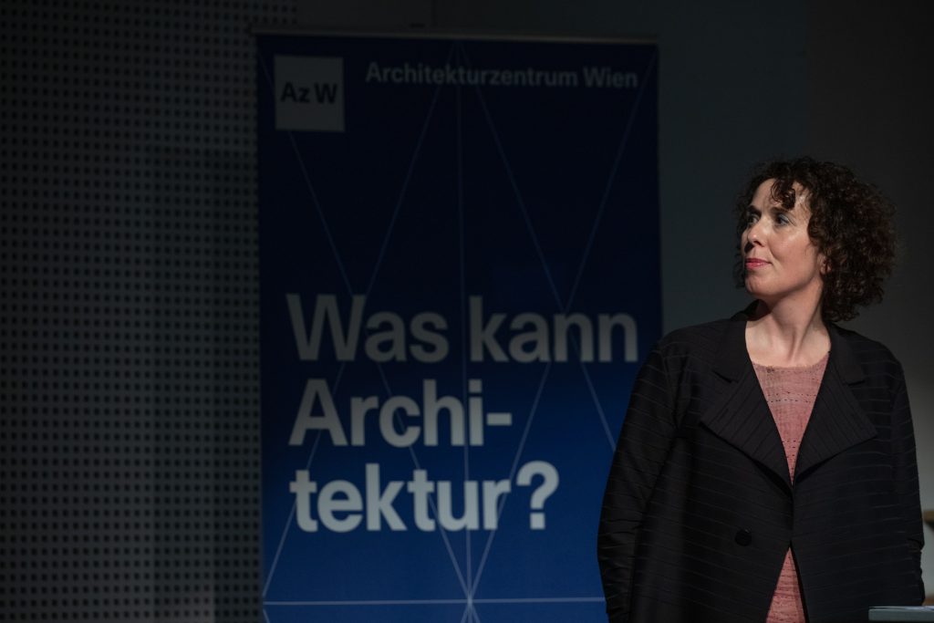 eine Frau mit dunklen gelockten Haaren schaut nach rechts, dahinter ist ein Schriftzug zu sehen
