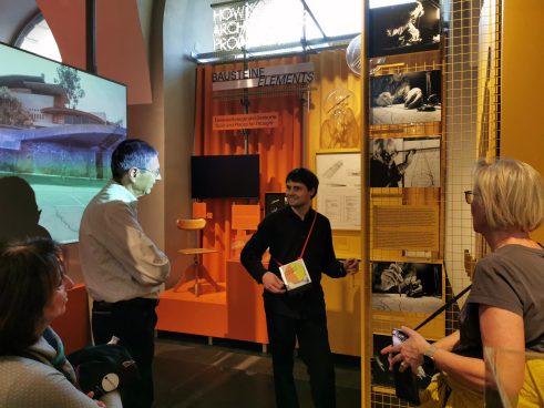 A man is standing in front of a group of people and explaining something. There are many objects, photos, and a screen in the room.
