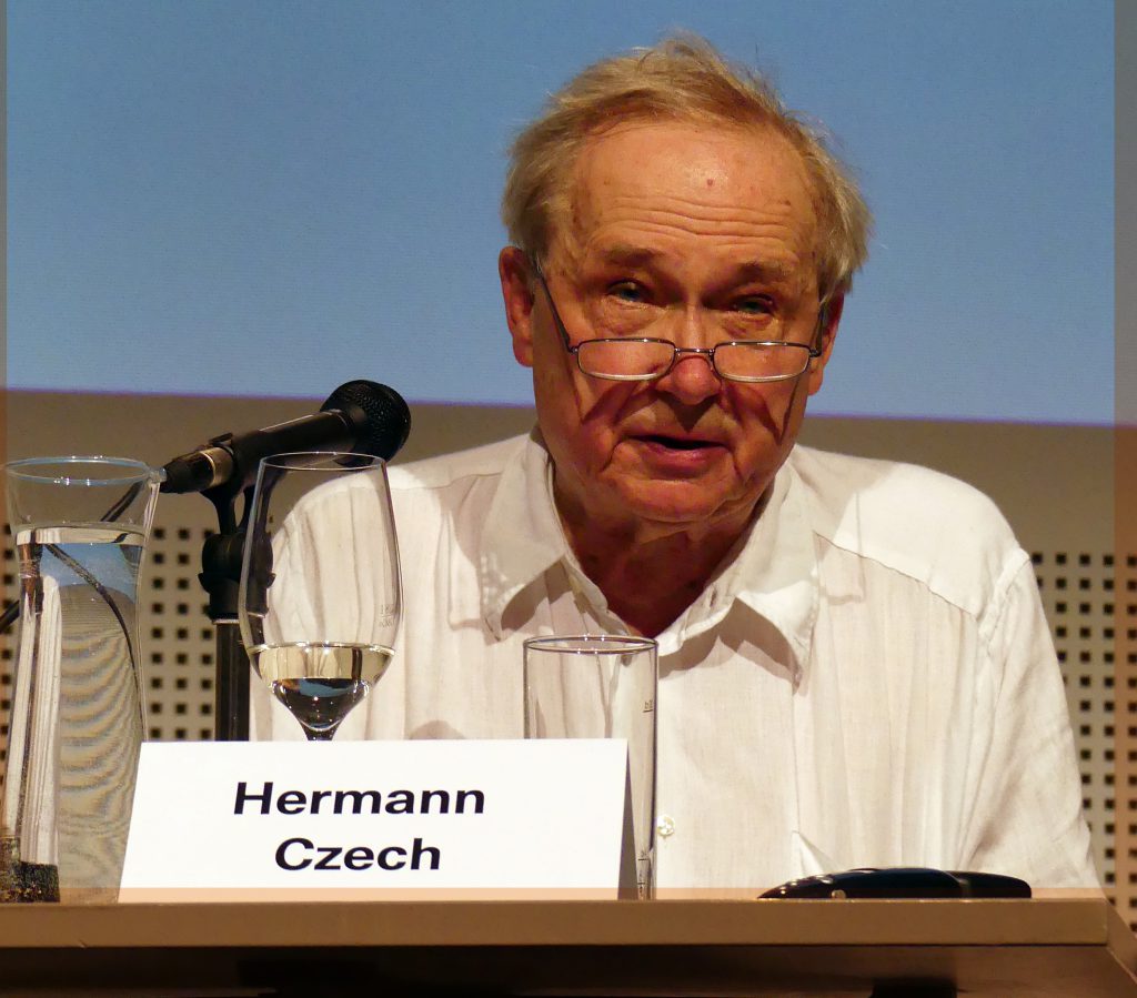 ein älterer Herr mit Brille und weißem Hemd sitz auf einem Tisch mit Mikrofon, vor ihm steht ein Schild worauf der Name Hermann Czech steht.