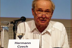 ein älterer Herr mit Brille und weißem Hemd sitz auf einem Tisch mit Mikrofon, vor ihm steht ein Schild worauf der Name Hermann Czech steht.