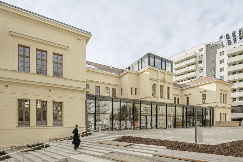 ein helles zweigeschossiges Gebäude mit einer flachen Treppe davor. Eine Frau in schwarz gekleidet geht die Stiegen hinauf.