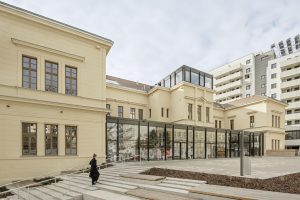 ein helles zweigeschossiges Gebäude mit einer flachen Treppe davor. Eine Frau in schwarz gekleidet geht die Stiegen hinauf.