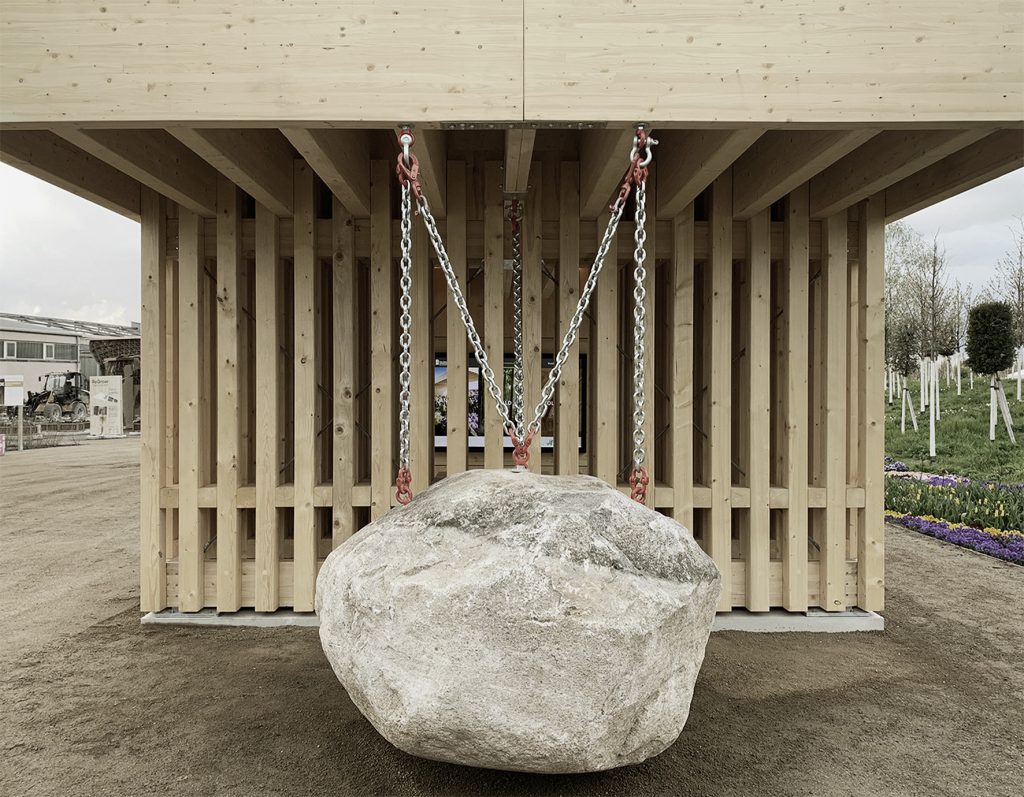 ein Holzbau mit vielen vertikalen Latten. Von oben hängt eine Metalkette an der ein riesiger Stein befestigt ist.