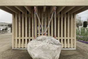 ein Holzbau mit vielen vertikalen Latten. Von oben hängt eine Metalkette an der ein riesiger Stein befestigt ist.
