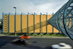 ein Gebäude in Gelb mit blauen Stelen und davor ist eine Straße mit vorbeifahrenden Autos
