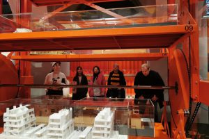 5 Personen stehen vor einem roten Gerüst auf dem sich mehrere Architekturmodelle befinden