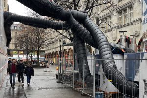 eine Straße auf der 3 Personen gehen, aus der Wand eines Hauses ragen schwarze dicke Schläuche zu einer Art Container.