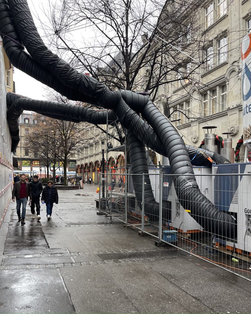eine Straße auf der 3 Personen gehen, aus der Wand eines Hauses ragen schwarze dicke Schläuche zu einer Art Container.