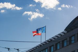 2 unterschiedliche Fahnen auf einem Dach eines Hauses. Die rechte Fahne ist blau mit gelben Sternen, die einen Kreis bilden und die linke Fahne ist rot weiß rot.